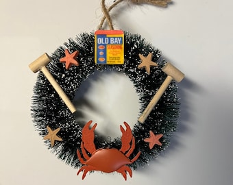 May include: A circular wreath with a dark green base, decorated with orange starfish, a crab, and wooden mallets. A small box of Old Bay Seasoning is at the top. The wreath is suspended by a twine loop.