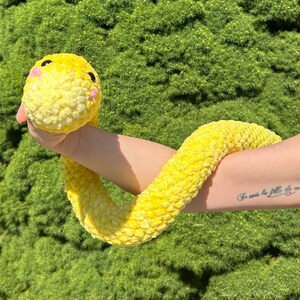 May include: A yellow crocheted snake toy with a pink nose and eyes. The snake is being held by a person's arm, which has a tattoo that reads "Je suis la fille de mes rêves".