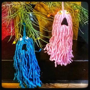 May include: Two yarn monsters with googly eyes and open mouths hanging from a pine branch. The monster on the left is blue and the monster on the right is pink.