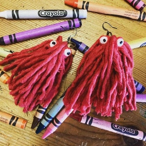 Puede incluir: Pendientes de monstruo de hilo rojo con ojos saltones blancos. Los pendientes se muestran con ceras Crayola de varios colores, incluyendo morado, naranja y blanco, sobre una superficie de madera.