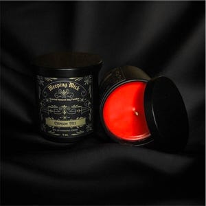 May include: A black candle jar with a gold label that reads "Weeping Wick Scented Natural Soy Candles Crimson Fizz The Irresistible Cherry 9 oz."