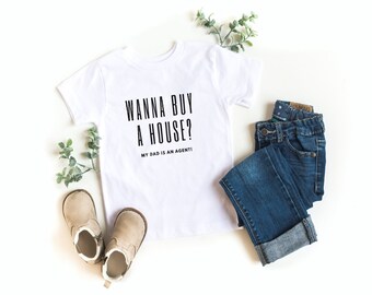 May include: White t-shirt with black text that reads "Wanna buy a house? My dad is an agent!" The shirt is laid flat on a white surface with a pair of brown boots and blue jeans.