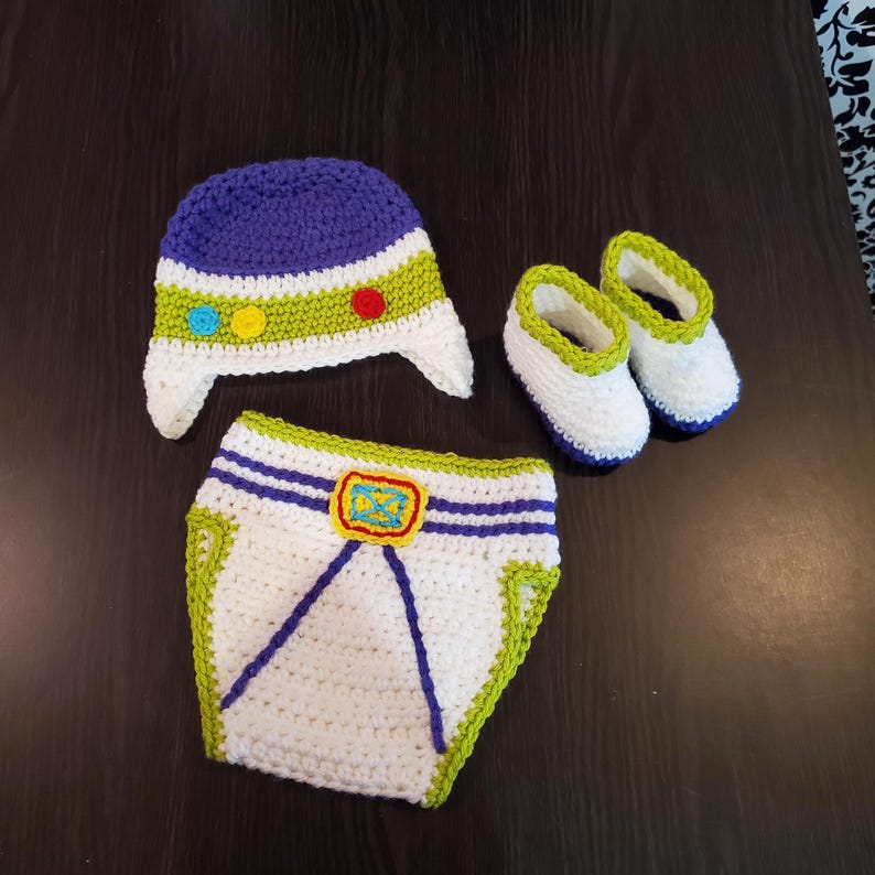 May include: A crochet baby outfit in white, blue, and green, featuring a hat, diaper cover, and booties. The hat has a blue top, white brim, and green stripe with colorful dots. The diaper cover has blue stripes and a yellow emblem. The booties have blue soles.