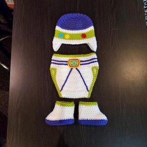 May include: A handmade crochet baby outfit in a space ranger theme. The set includes a blue and white hat, diaper cover, and booties. The hat has a green and white band with colorful accents. The diaper cover has a yellow buckle.