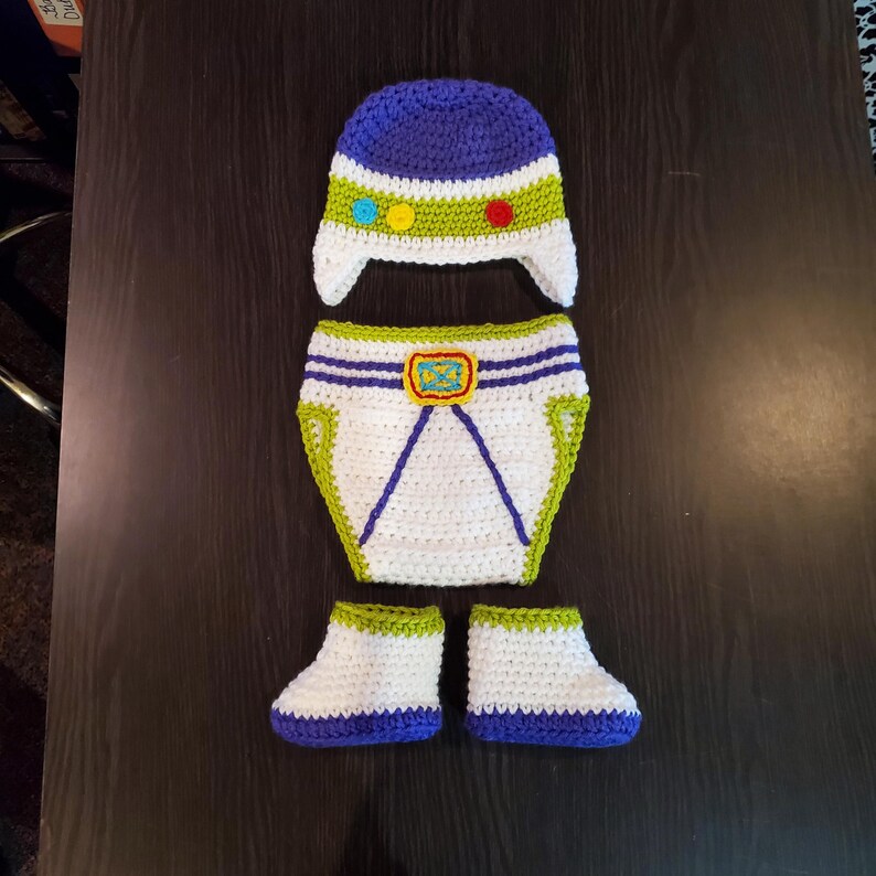 May include: A handmade crochet baby outfit in a space ranger theme. The set includes a blue and white hat, diaper cover, and booties. The hat has a green and white band with colorful accents. The diaper cover has a yellow buckle.