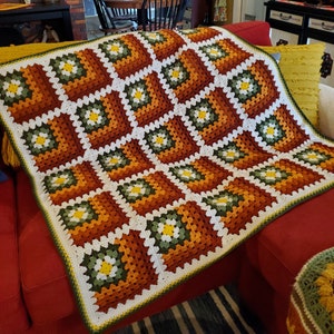 May include: A crocheted blanket with a geometric pattern in shades of brown, green, white, and yellow. The blanket is made of granny squares with a flower design in the center of each square.