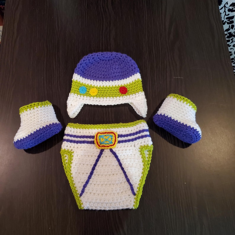 May include: A handmade crochet baby outfit in a space theme. The set includes a white and blue hat, diaper cover, and two booties. The hat has a green band with colorful buttons. The diaper cover has a yellow buckle.