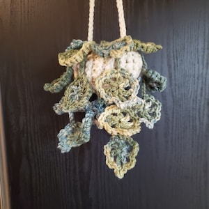 May include: A crocheted hanging planter with a white base and green and blue leaves. The planter is hanging from a white cord.