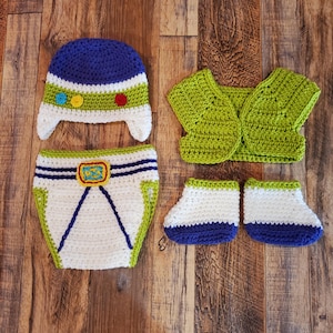 May include: A handmade crochet baby outfit in a space ranger theme. The set includes a blue and white hat, a pair of booties, a diaper cover, and a vest. The outfit features green, white, blue, and yellow accents.