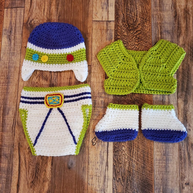 May include: A handmade crochet baby outfit in a space ranger theme. The set includes a blue and white hat, a pair of booties, a diaper cover, and a vest. The outfit features green, white, blue, and yellow accents.
