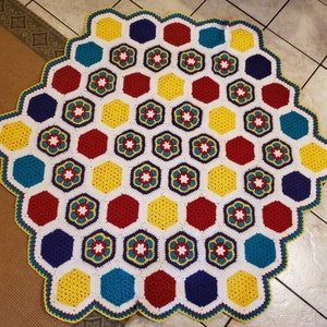 May include: A crocheted hexagonal blanket with a white background and colorful hexagons in red, yellow, blue, and purple. The blanket has a scalloped edge.