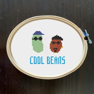 May include: A white embroidery hoop with a cross-stitch design of two cartoon beans wearing sunglasses and hats. The text "COOL BEANS" is stitched in blue below the beans.
