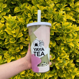 May include: A green and white reusable cup with a straw. The cup has a pink and white design with a cartoon character on it. The text "YAYA TEA" is printed on the cup.