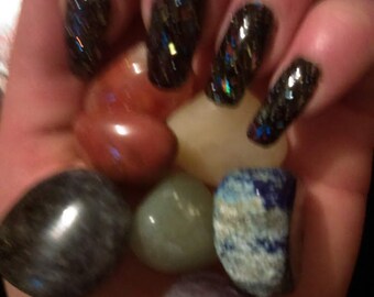 May include: A hand holding a variety of smooth, polished stones in different colours, including red, green, blue, and purple. The hand is also wearing black nail polish with glitter.