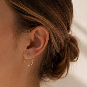 May include: A gold-toned earring featuring a small spherical stud at the top of the earlobe and a thin, circular hoop suspended from the back, creating a double-layered effect that sits flush against the lower part of the earlobe.