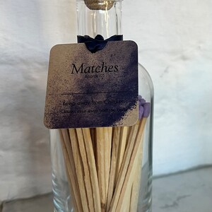May include: A glass bottle filled with approximately 125 wooden matches with purple tips. The bottle has a wooden cork stopper and a brown paper label that reads "Matches Approx 125 Keep away from Children Always strike away from the body".