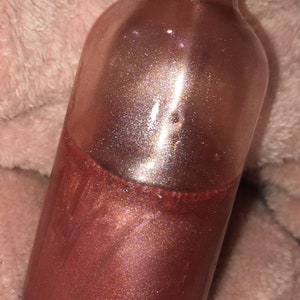 May include: A clear glass bottle with a black cap, filled with a shimmering, bronze-colored liquid. The liquid has a glittery, iridescent effect. The bottle is partially filled, with the liquid taking up the lower half. The background is a soft, pinkish-beige fabric.