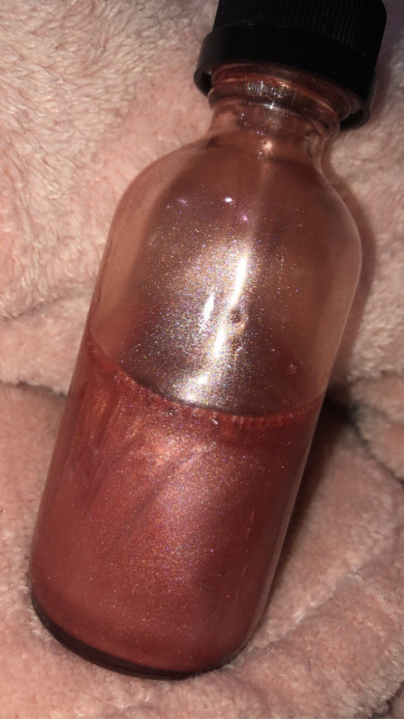 May include: A clear glass bottle with a black cap, filled with a shimmering, bronze-colored liquid. The liquid has a glittery, iridescent effect. The bottle is partially filled, with the liquid taking up the lower half. The background is a soft, pinkish-beige fabric.