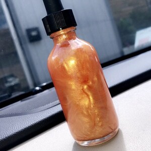 May include: A clear glass bottle filled with a shimmering, golden-orange liquid. The bottle has a black dropper top. The product is displayed on a white surface, with a dark dashboard visible in the background.