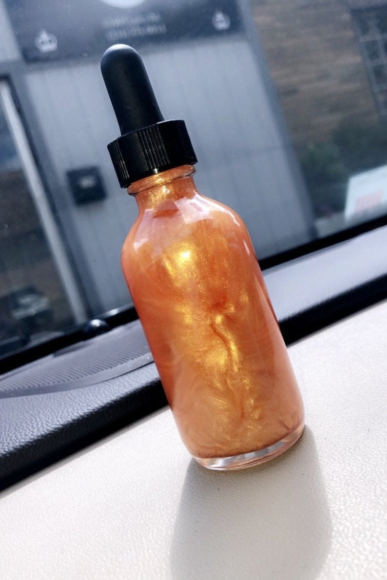 May include: A clear glass bottle filled with a shimmering, golden-orange liquid. The bottle has a black dropper top. The product is displayed on a white surface, with a dark dashboard visible in the background.