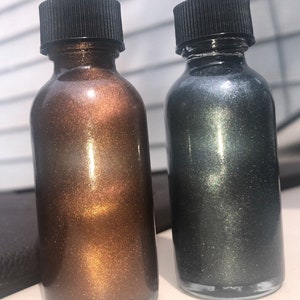 May include: Two small glass bottles with black caps. One bottle contains a bronze-colored liquid with a shimmering effect, and the other contains a dark gray liquid with a similar shimmer. The bottles are side-by-side.