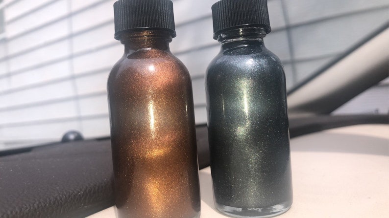 May include: Two small glass bottles with black caps. One bottle contains a bronze-colored liquid with a shimmering effect, and the other contains a dark gray liquid with a similar shimmer. The bottles are side-by-side.