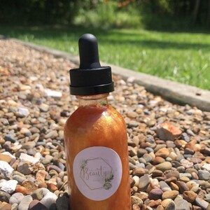May include: A clear glass bottle with a black dropper top, filled with a shimmering, golden-orange liquid. A white label with the text "Beauty" and a botanical design is affixed to the bottle. The bottle is set on a bed of small, multicolored stones.
