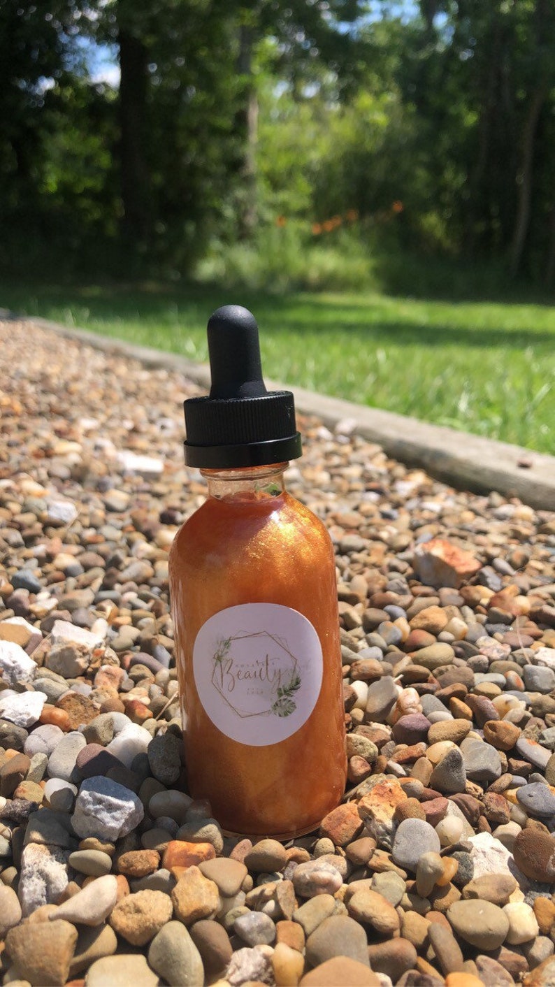May include: A clear glass bottle with a black dropper top, filled with a shimmering, golden-orange liquid. A white label with the text "Beauty" and a botanical design is affixed to the bottle. The bottle is set on a bed of small, multicolored stones.