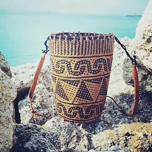May include: A woven brown and black basket with a geometric pattern. The basket has a long strap and is sitting on a rocky surface.