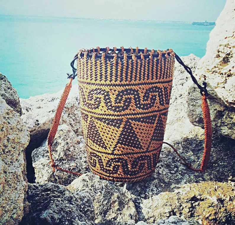 May include: A woven brown and black basket with a geometric pattern. The basket has a long strap and is sitting on a rocky surface.