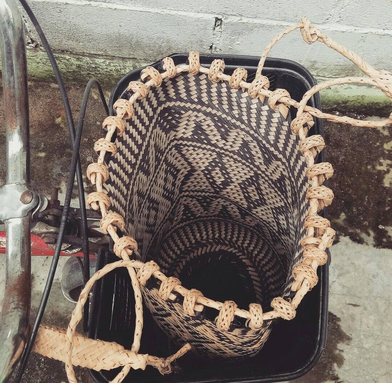 May include: A woven basket with a black and white pattern is attached to a bicycle. The basket has a brown leather handle and is made of natural materials.
