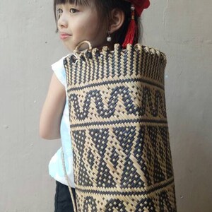 May include: A young girl wearing a white shirt and black shorts is carrying a woven black and brown bag with a geometric pattern. The bag has a strap that goes over the shoulder. The girl is wearing a red bow in her hair.