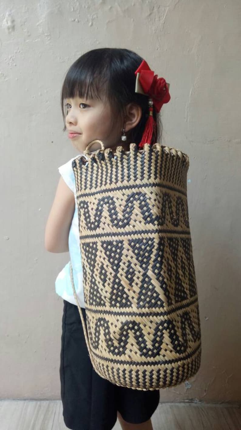 May include: A young girl wearing a white shirt and black shorts is carrying a woven black and brown bag with a geometric pattern. The bag has a strap that goes over the shoulder. The girl is wearing a red bow in her hair.