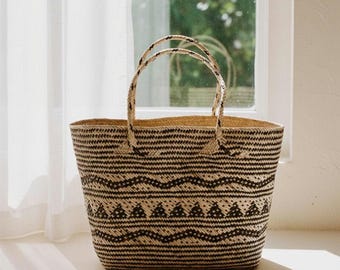 May include: A woven straw tote bag featuring a repeating pattern of horizontal black stripes, wavy lines, and triangular shapes. The bag has two matching woven straw handles attached to the top rim, creating a structured open-top carryall design.