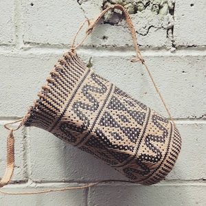 May include: A woven bag with a black and brown geometric pattern. The bag has a long strap and is hanging from a white brick wall.