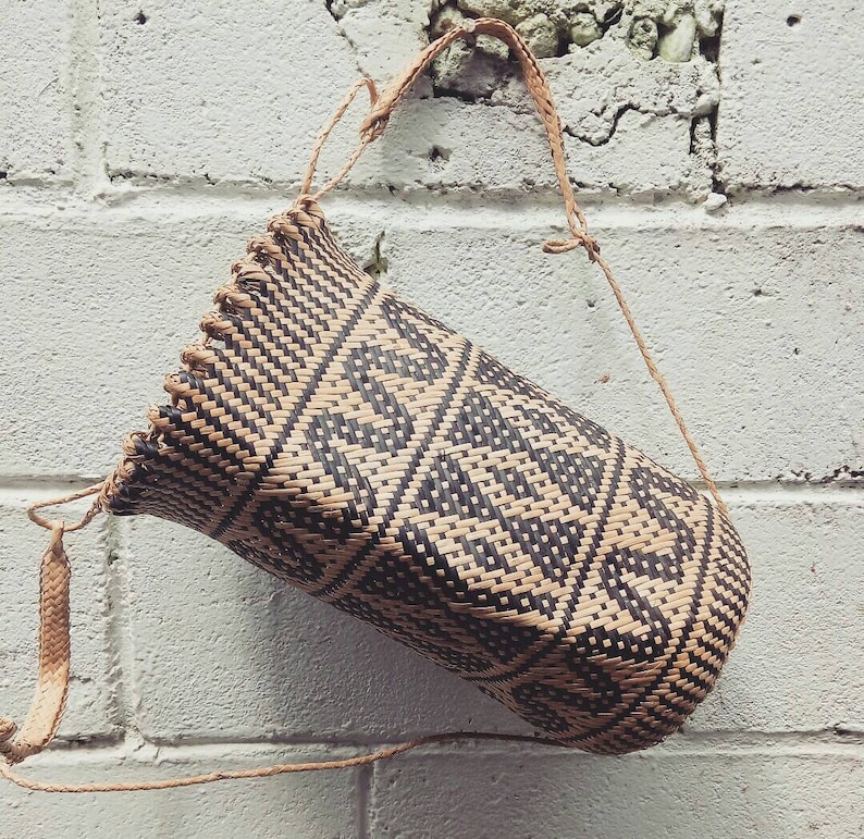 May include: A woven bag with a black and brown geometric pattern. The bag has a long strap and is hanging from a white brick wall.