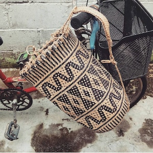 May include: A black and tan woven basket bag with a geometric pattern hangs from a bicycle.