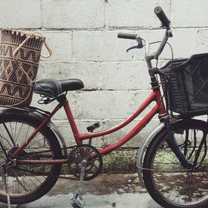 May include: A red bicycle with a black basket and a woven bag on the back rack. The bicycle is parked in front of a white brick wall.