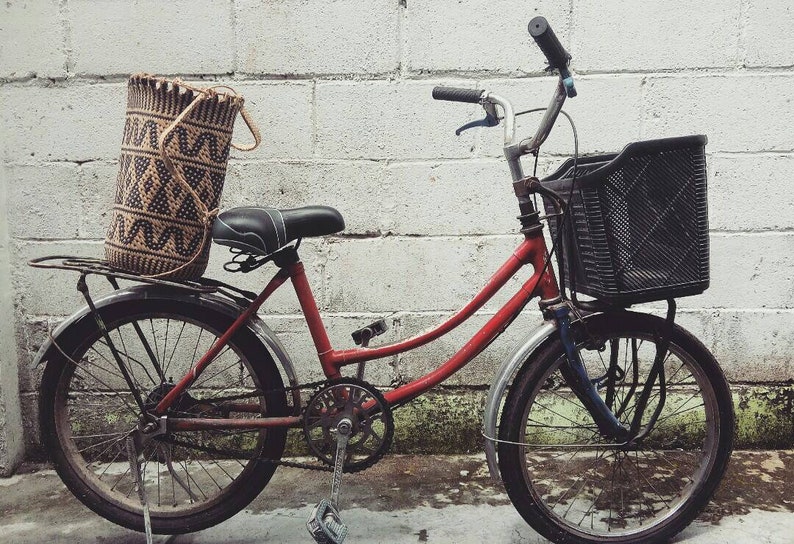 May include: A red bicycle with a black basket and a woven bag on the back rack. The bicycle is parked in front of a white brick wall.