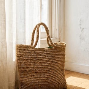 May include: A light brown woven tote bag with two handles. The bag is rectangular in shape. The bag is on a wooden floor next to a window with white curtains. The bag is made of a natural material.