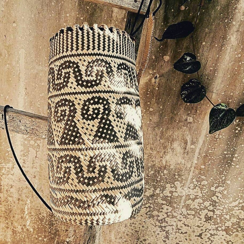 May include: A woven cylindrical basket with a black and white geometric pattern. The basket is hanging from a hook on a wooden beam.