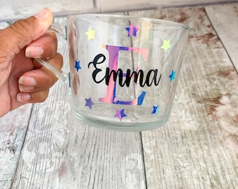May include: Clear glass mug with a holographic iridescent letter "E" and the name "Emma" in black lettering. The mug is decorated with yellow and blue stars.