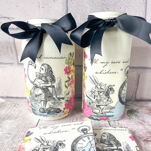 Op de afbeelding: Twee witte potten met zwarte strikken en Alice in Wonderland-decoupage. De potten bevatten citaten uit het boek: "Oh my ears and whiskers." en "And curiouser." De potten staan op een witte bakstenen achtergrond.