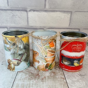 Puede incluir: Tres latas de conserva recicladas decoradas con decoupage con temas de animales y naturaleza. Las latas están adornadas con imágenes de un burro, una ardilla y pájaros, atadas con hilo. Las latas están dispuestas en fila.