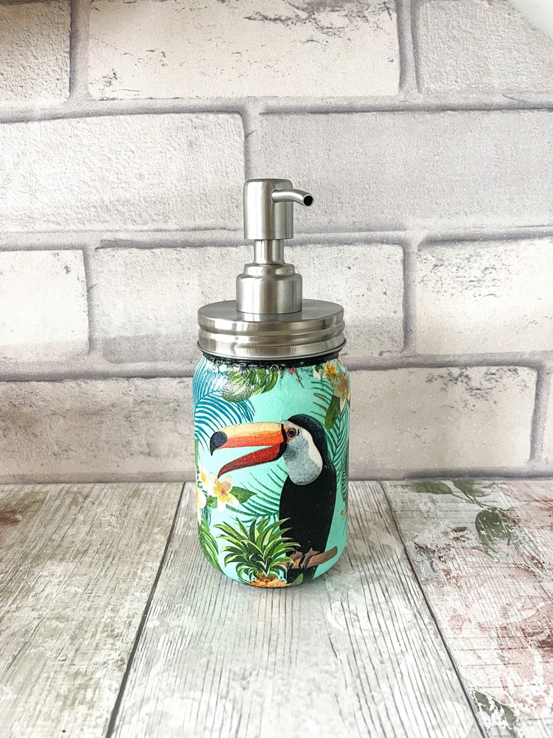 May include: A turquoise glass soap dispenser with a stainless steel pump and a tropical toucan design. The dispenser is decorated with green palm leaves, yellow flowers, and a pineapple.
