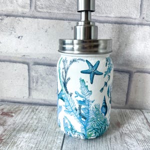 May include: A white ceramic soap dispenser with a stainless steel pump and lid. The dispenser is decorated with a blue and green sea life pattern, including starfish, seaweed, and seashells.