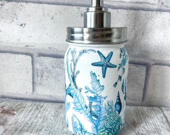 May include: A white ceramic soap dispenser with a stainless steel pump and lid. The dispenser is decorated with a blue and green sea life pattern, including starfish, seaweed, and seashells.