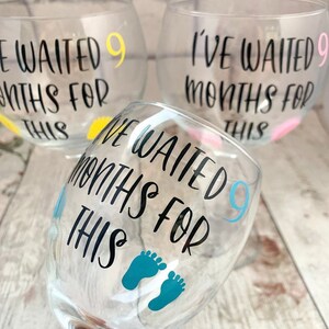 May include: Three clear glass wine glasses with the text "I've waited 9 months for this" on each. The glasses have different colored accents: yellow, pink, and blue. The blue glass has a pair of blue footprints.