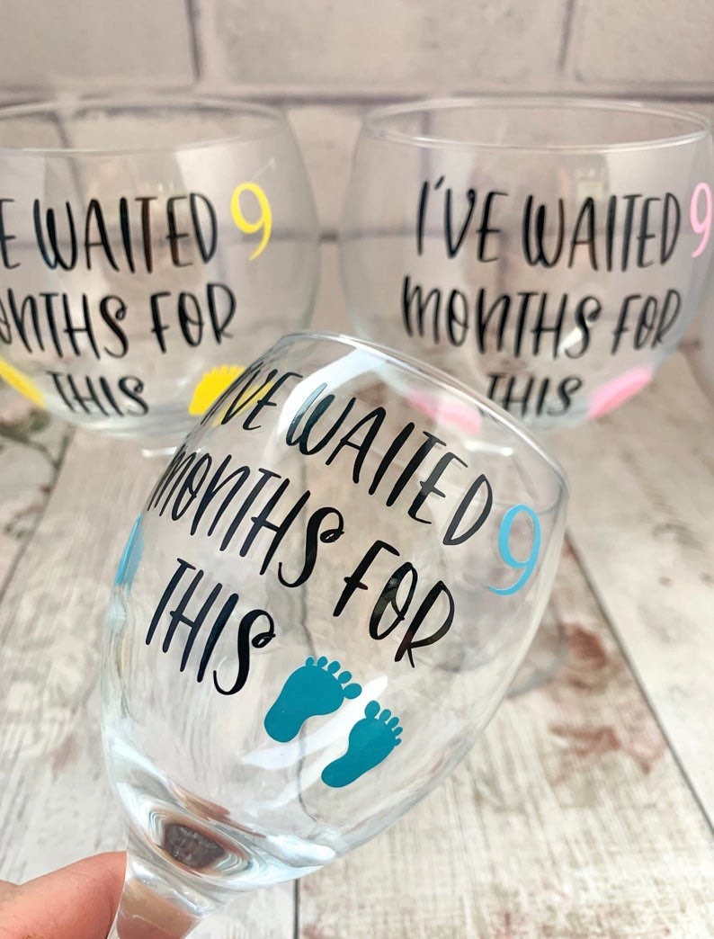 May include: Three clear glass wine glasses with the text "I've waited 9 months for this" on each. The glasses have different colored accents: yellow, pink, and blue. The blue glass has a pair of blue footprints.