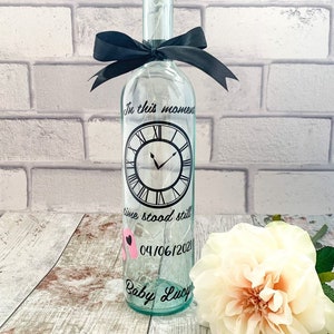 May include: A clear glass bottle with a cork top and a black ribbon tied around the neck. The bottle has a clock face with the words "In this moment time stood still" and the date "04/06/2021". The bottle also has the words "Baby Lucy" written on it.